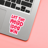 Laptop with a sticker reading 'Let the Weird Idea Win' on a pink surface